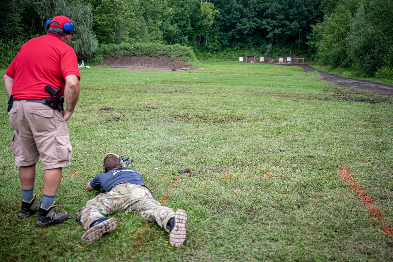A soldier laying on the ground fires a weapon as a person watches.