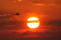 A helicopter flies in an orangish sky as the sun sits to the right.