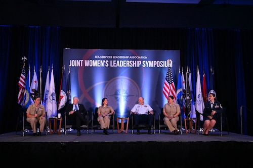 photo of a stage with men and women, civilian and U.S. military, sitting in chairs with a projector showing Joint Women's Leadership Symposium