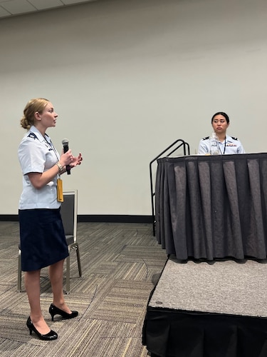 photo of two uniformed U.S, Space Force Guardians, one standing holding a microphone, one sitting at a table on a stage