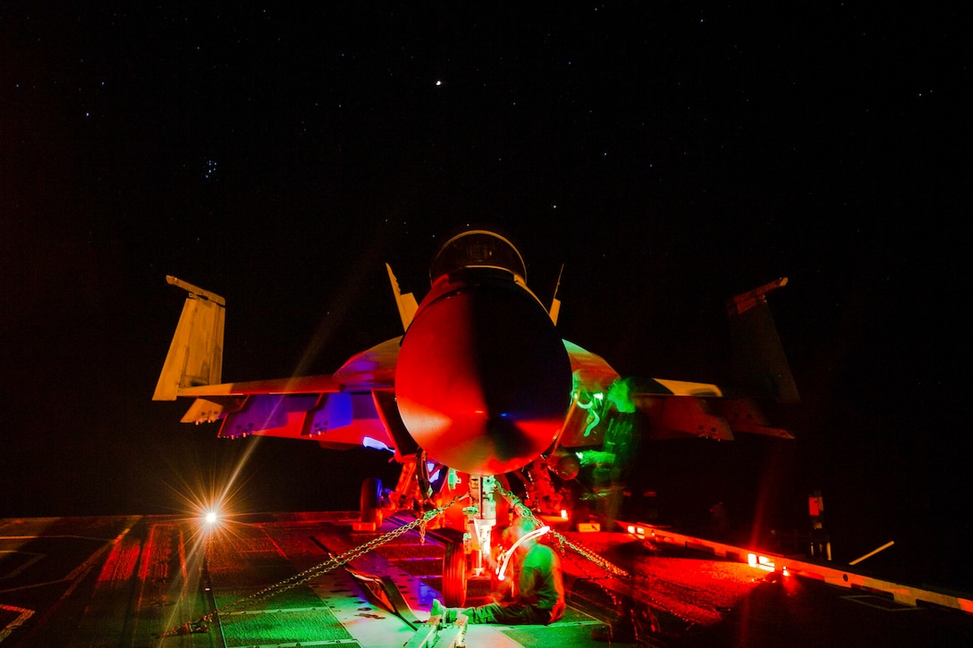 An aircraft illuminated by red and green lights parked on the deck of a ship at night.