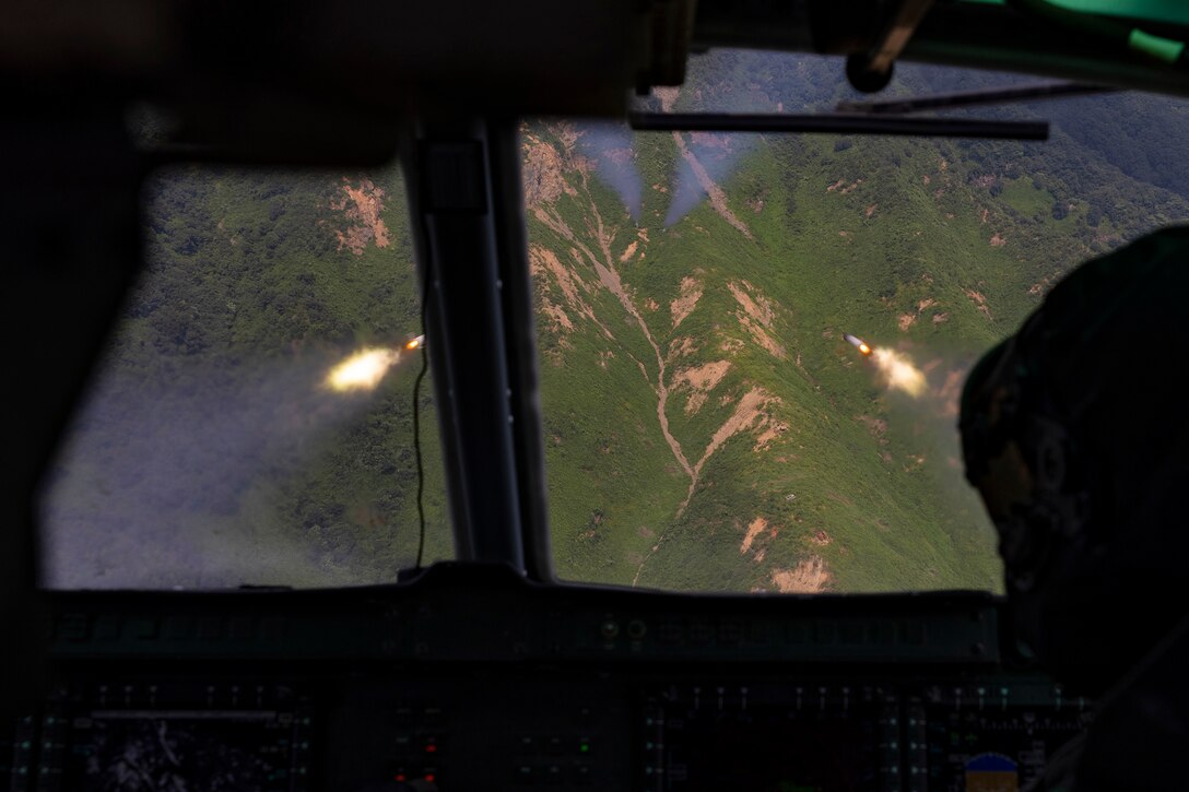 Marines watch from the cockpit of a military aircraft as rockets fire over mountainous terrain.