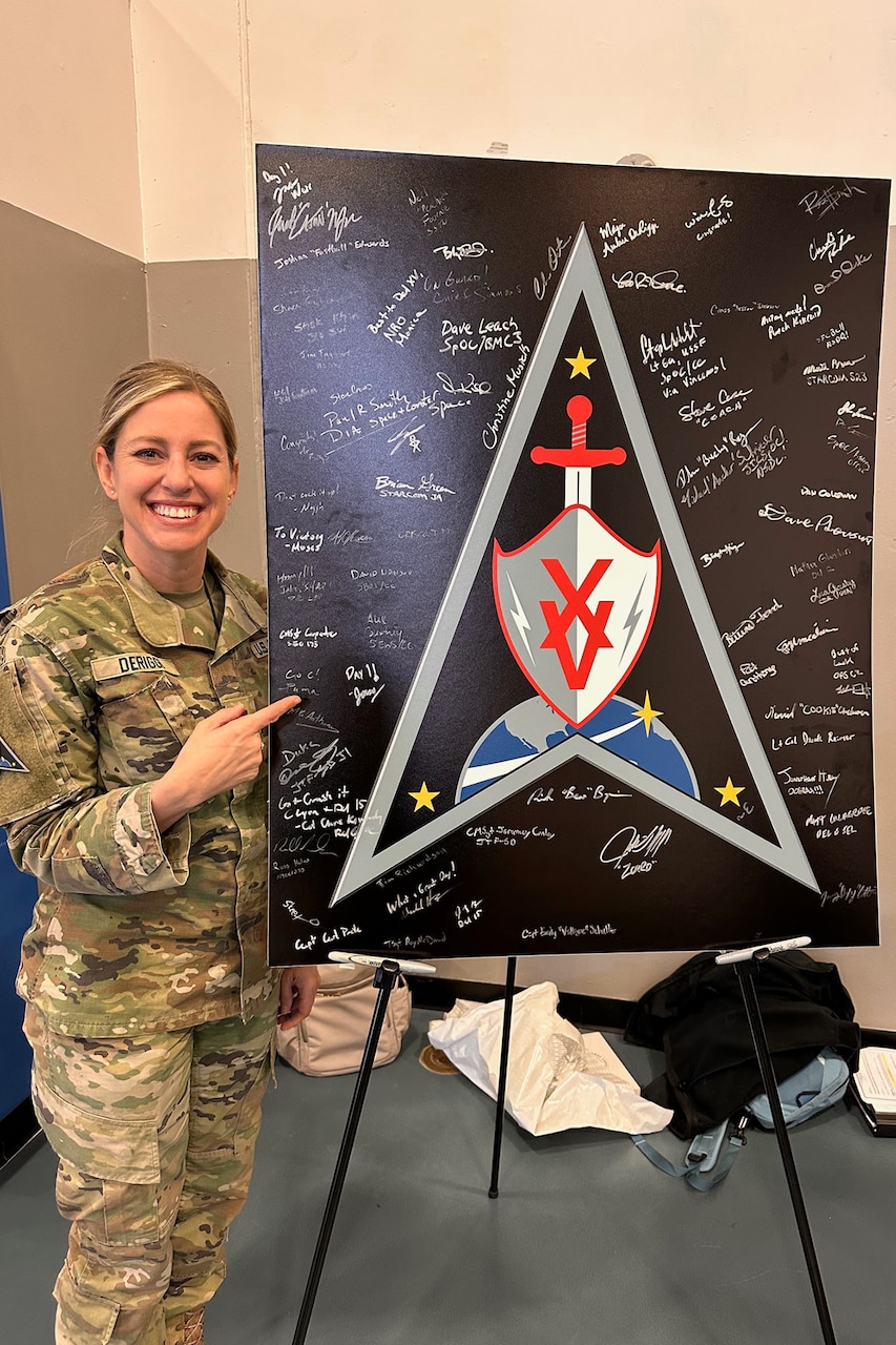 A uniformed service member stands next to a sign with a logo containing multiple signatures.