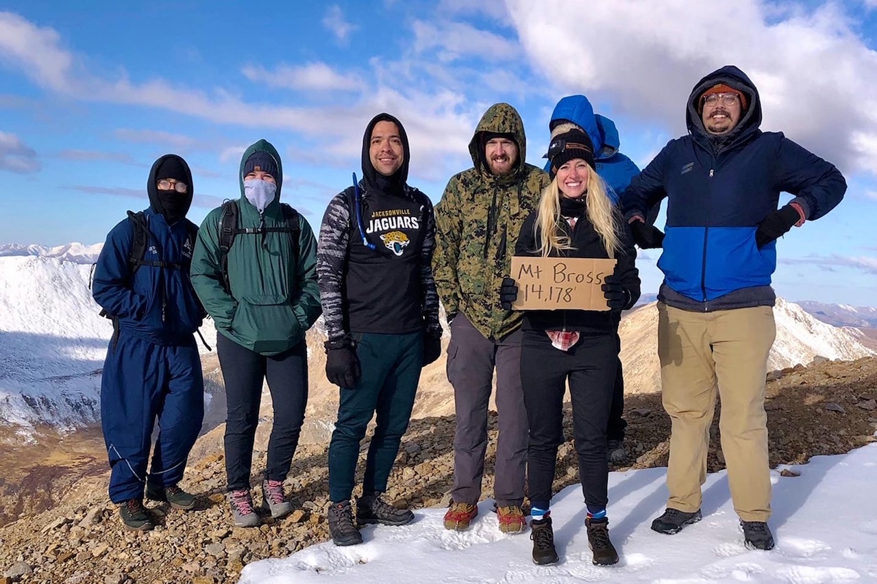 A group of people, wearing cold weather gear, stand atop a snowy mountain.