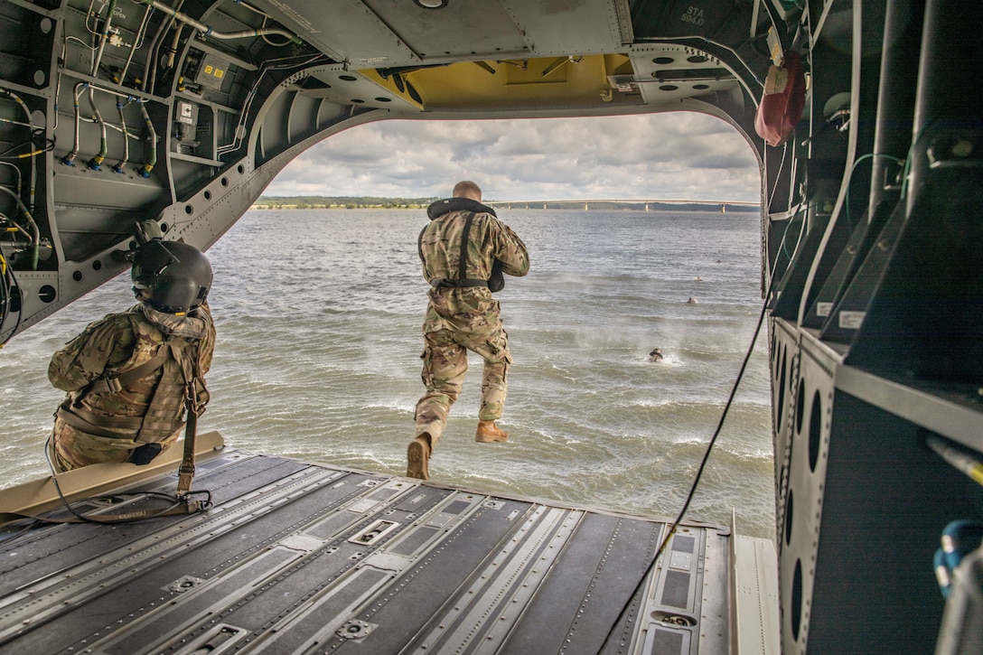 A soldier wearing a life vest jumps into a body of water from an aircraft as a fellow service member sits to the left on the open doorway.