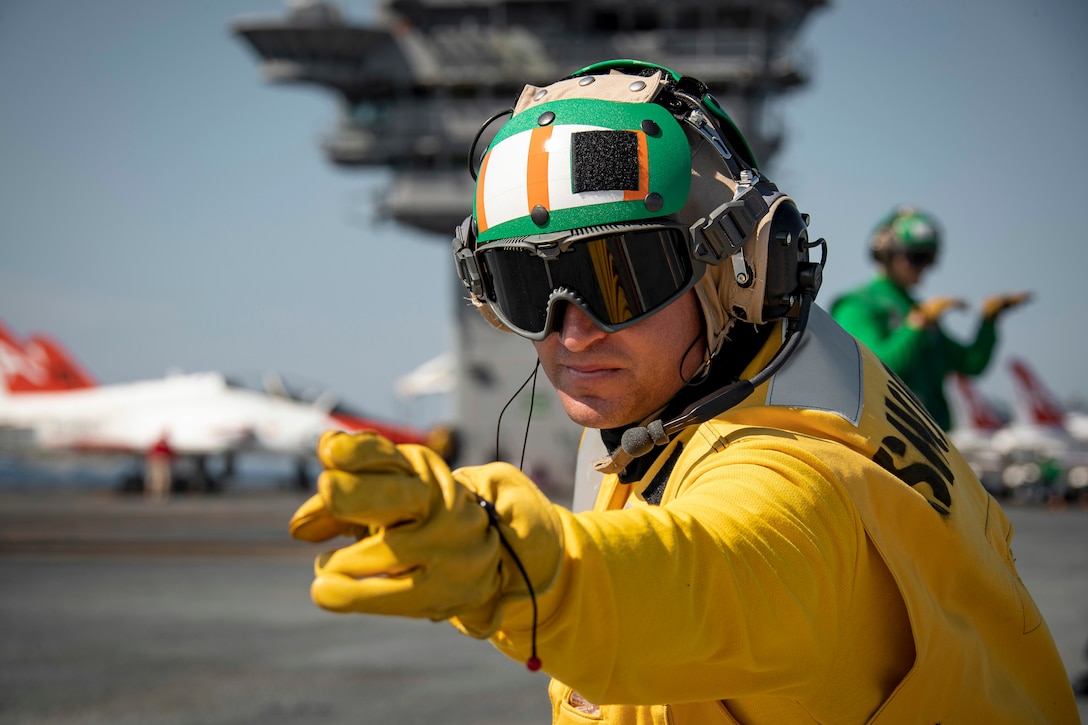 A closeup of a sailor wearing protective gear while pointing with blurred aircraft in the background.