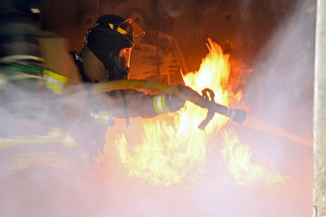 An airman wearing firefighter gear uses a hose to spray water on a fire as smoke covers the foreground.