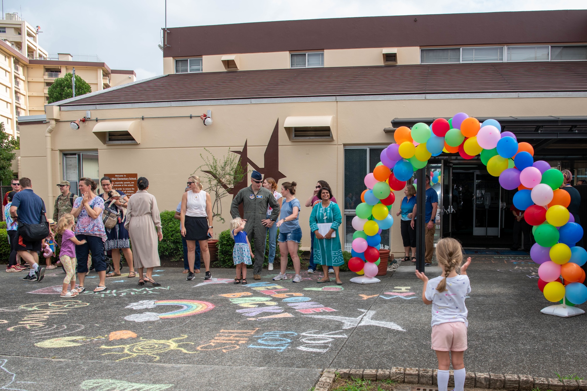 Parents, faculty, and community members stand outside of a school to welcome students inside.
