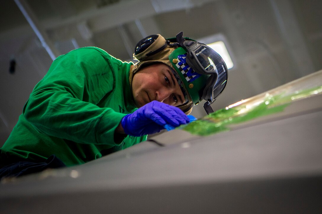 A sailor wearing a disposable glove leans forward and uses a smoothing tool on a military aircraft.