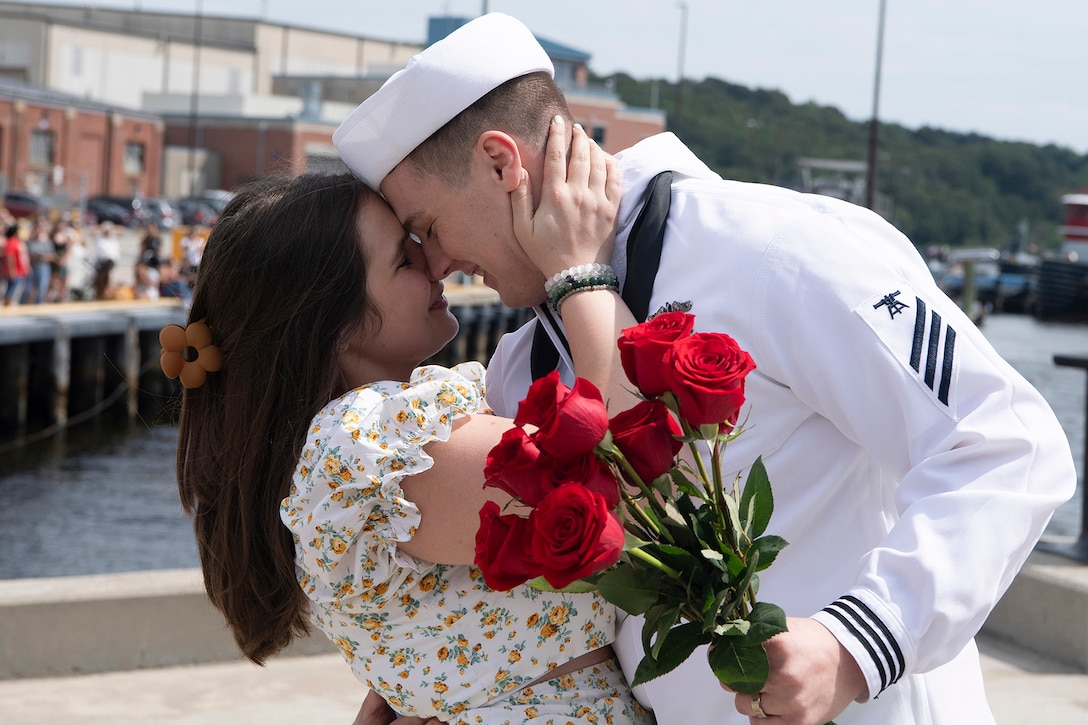 A uniformed sailor holding roses embraces a smiling partner.