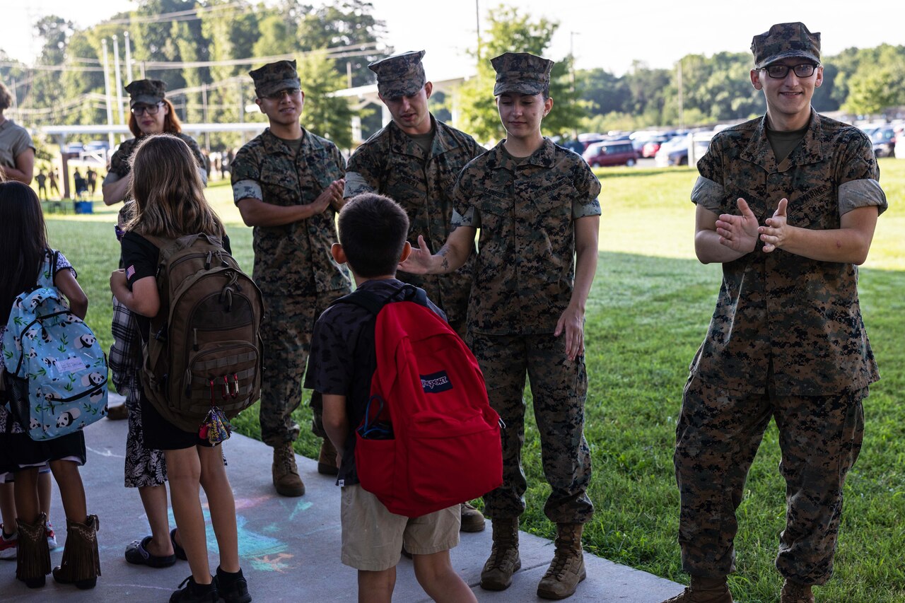 Six Marines line up and greet three children walking past wearing backpacks.