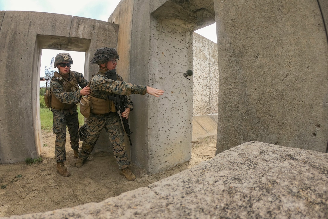 A Marine holds a fellow Marine throwing a grenade in an open concrete structure.