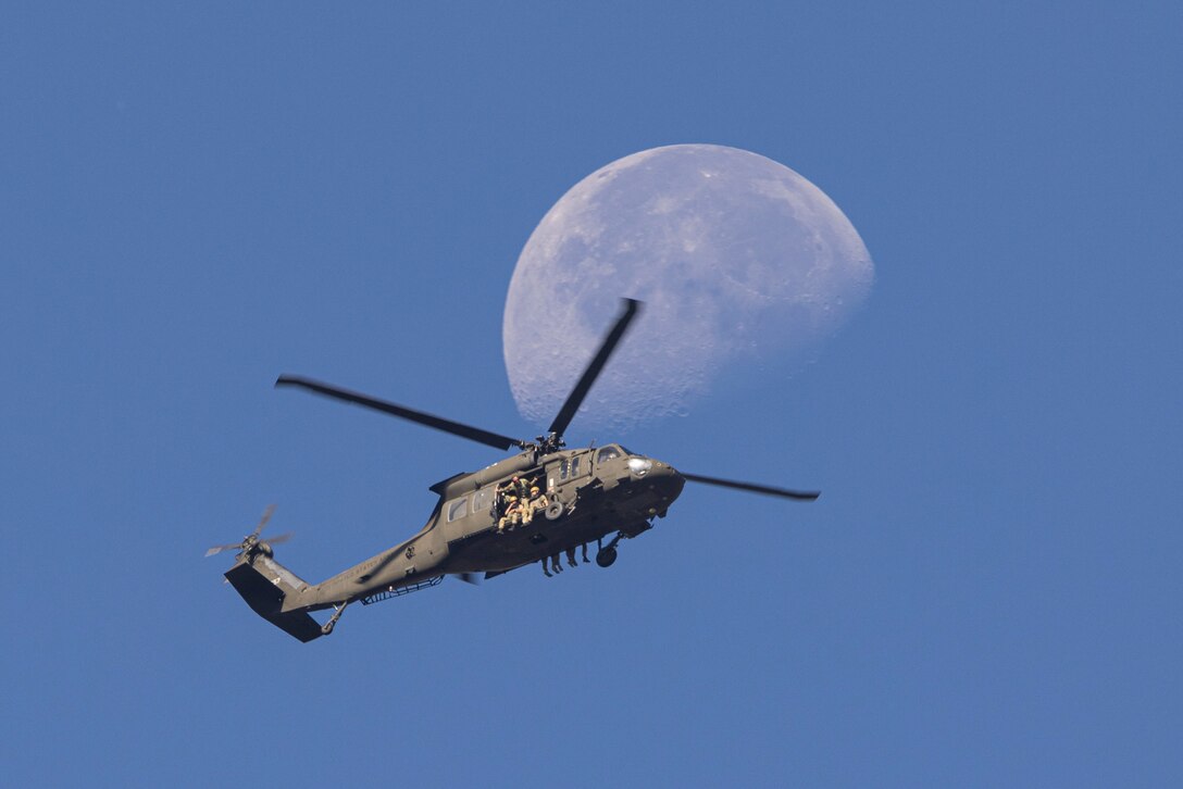 Soldiers sit in the open doorway of an airborne helicopter with the moon in the background.