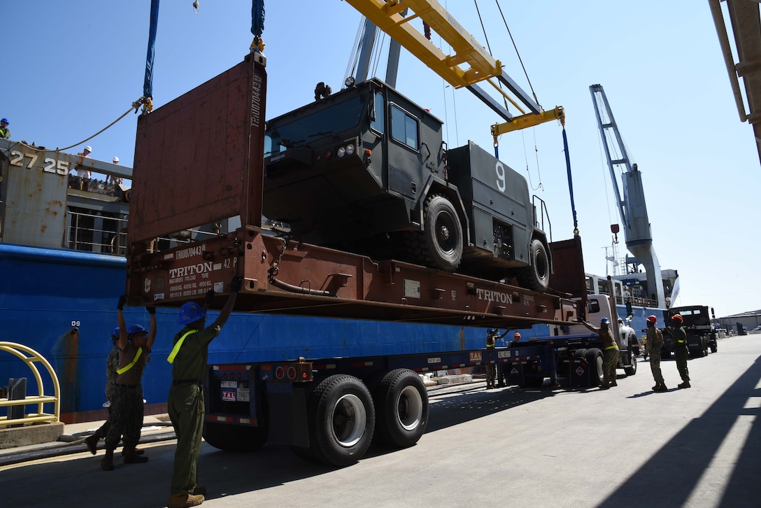 Photo shows sailors guiding a military fire truck onto a truck flat bed on a pier.