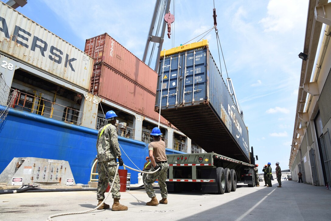 Photo shows two sailors in the foreground guiding a large storage container onto a truck flatbed.