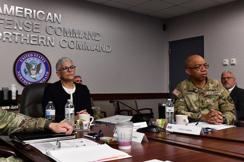 A woman in civilian attire sits at a table with others who are in military uniforms.