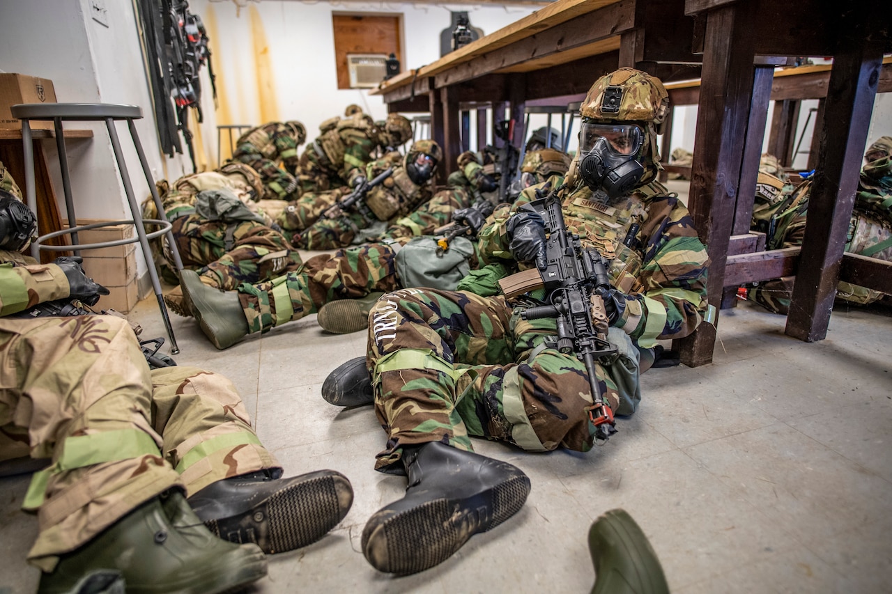 Service members wearing gas masks sit on a floor during an exercise.