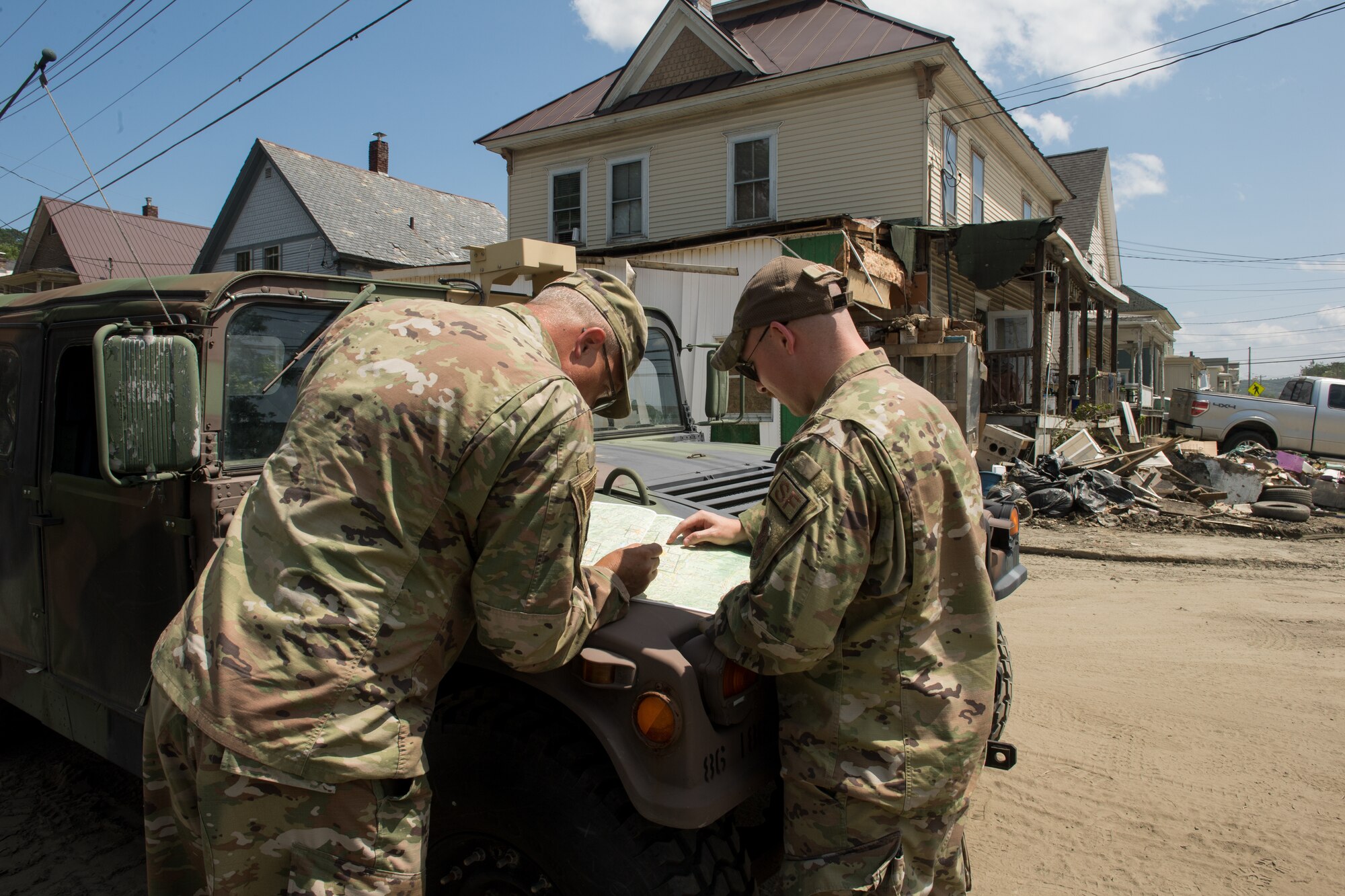 Photo of Senior Master Sgt. Matthew Powell (left), 158th Cyber Operations superintendent, and Tech. Sgt. Brandon Matott, 158th Security Forces Squadron personnel, surveying a map as part of a Liaison Officer Mission, amidst rubble and debris caused by historic flooding in Barre, Vermont, July 19, 2023. The purpose of the Liaison Officer mission is to meet with town leaders and report information from the town back to the state for possible resourcing. (U.S. Air Force photo by Tech. Sgt. Richard Mekkri)