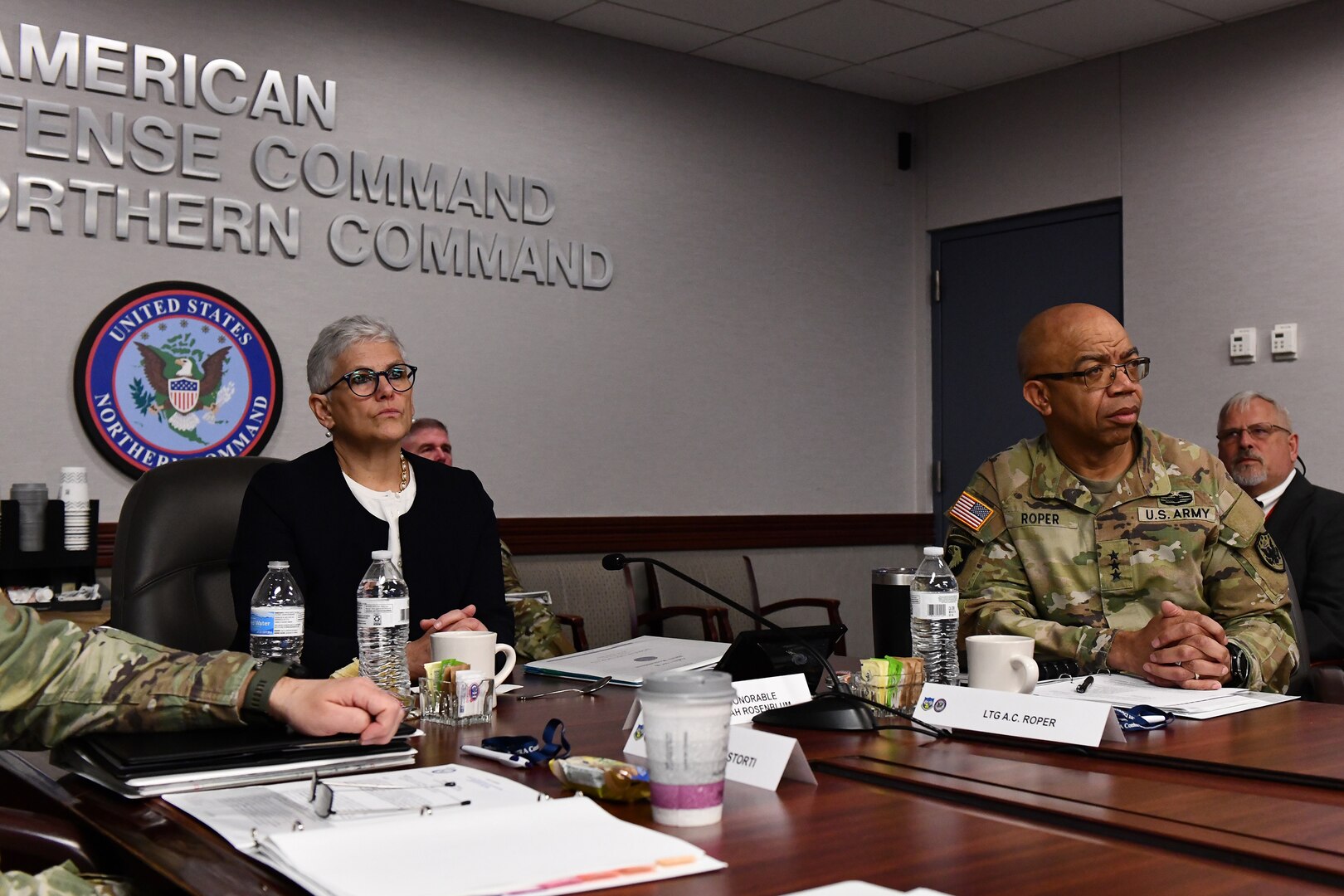 A woman in civilian attire sits at a table with others who are in military uniforms.