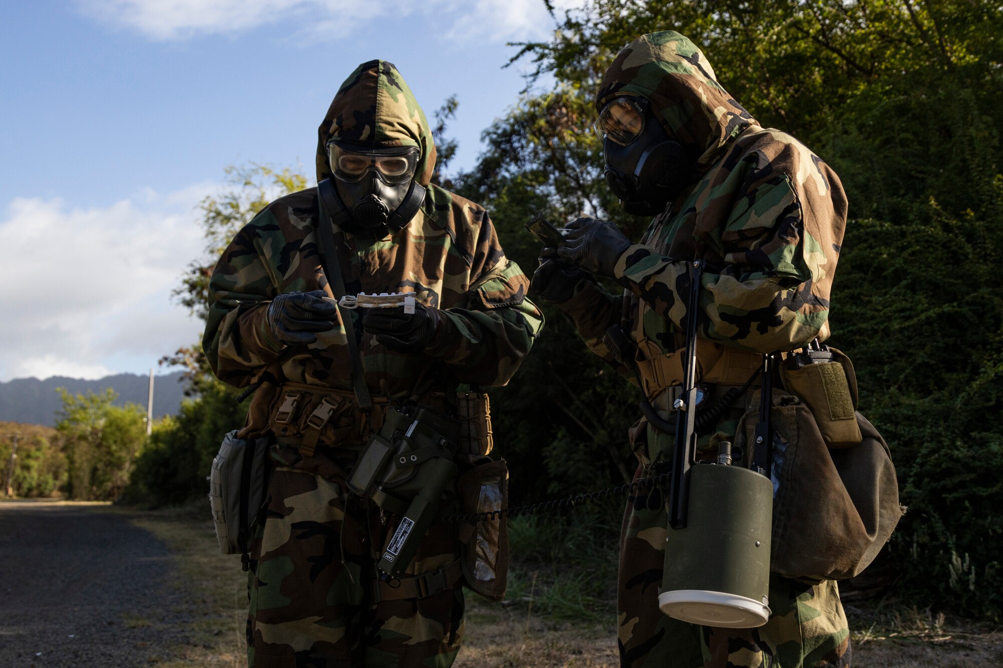 Service members wearing gas masks and protective gear stand outdoors during an exercise.