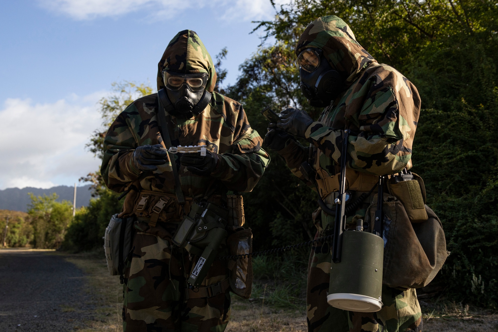 Service members wearing gas masks and protective gear stand outdoors during an exercise.