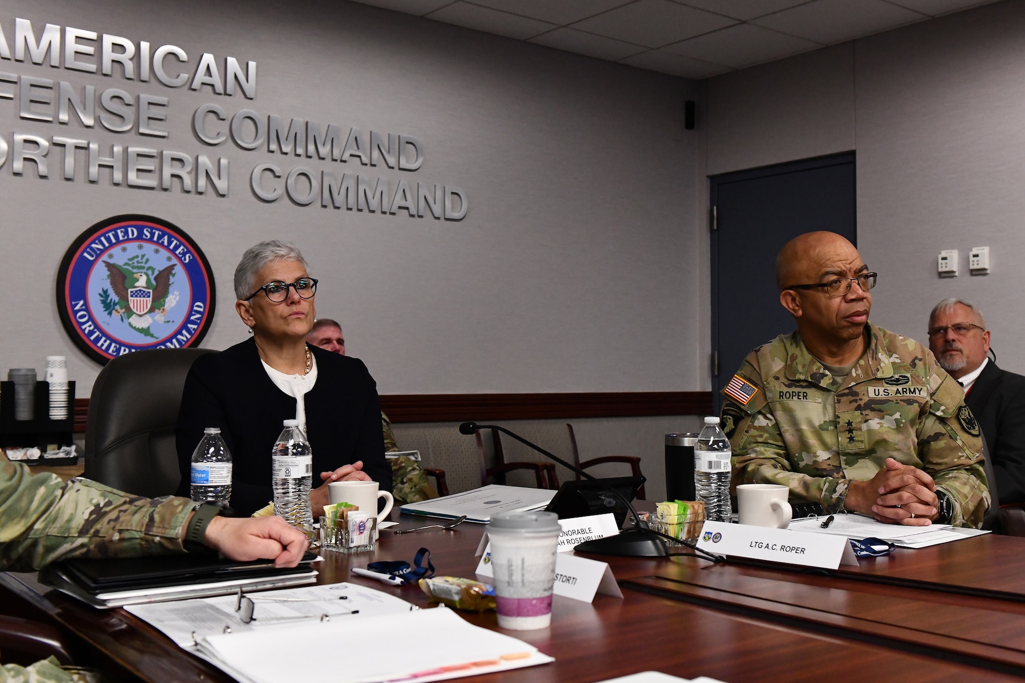 A woman in civilian attire sits at a table with others who are in military uniforms.