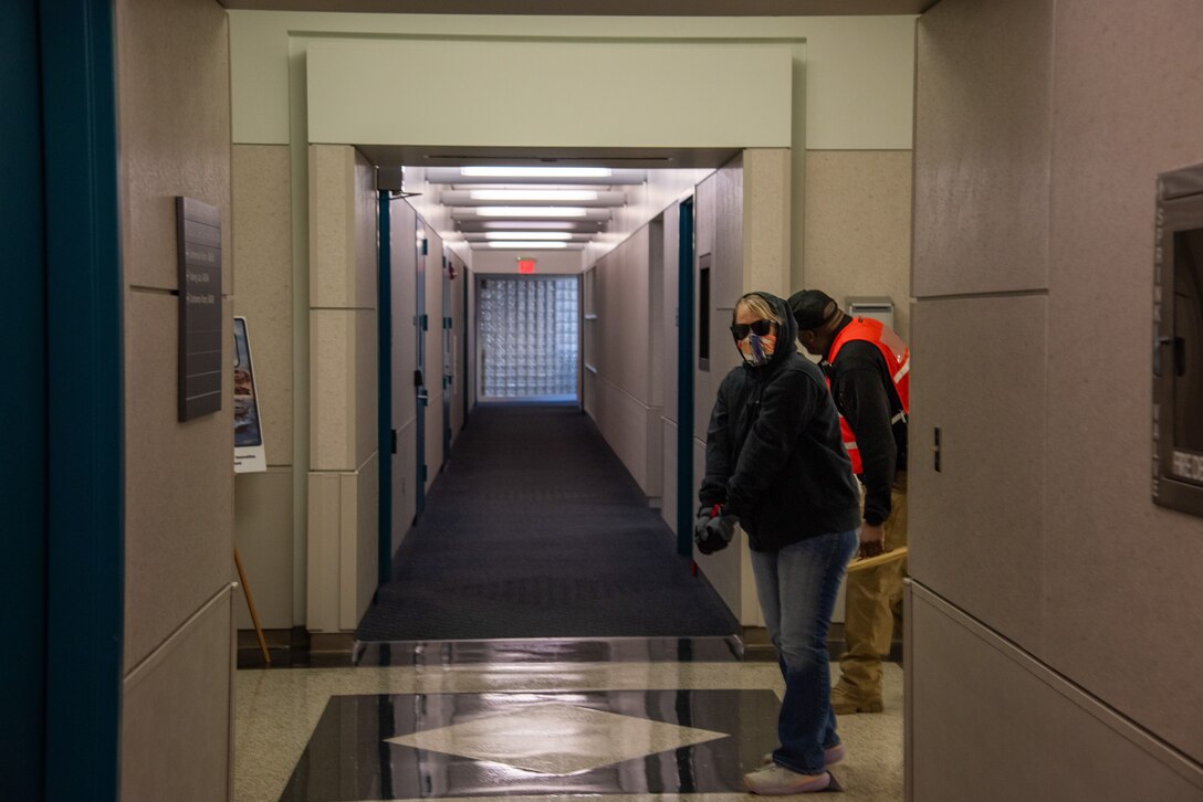 A woman wearing sunglasses, blue jeans, a blue hoodie and an orange and white mask coving nose and mouth, turns corner in a hallway. A man in orange vest looks the other way.