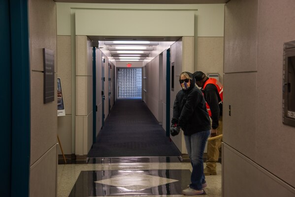 A woman wearing sunglasses, blue jeans, a blue hoodie and an orange and white mask coving nose and mouth, turns corner in a hallway. A man in orange vest looks the other way.