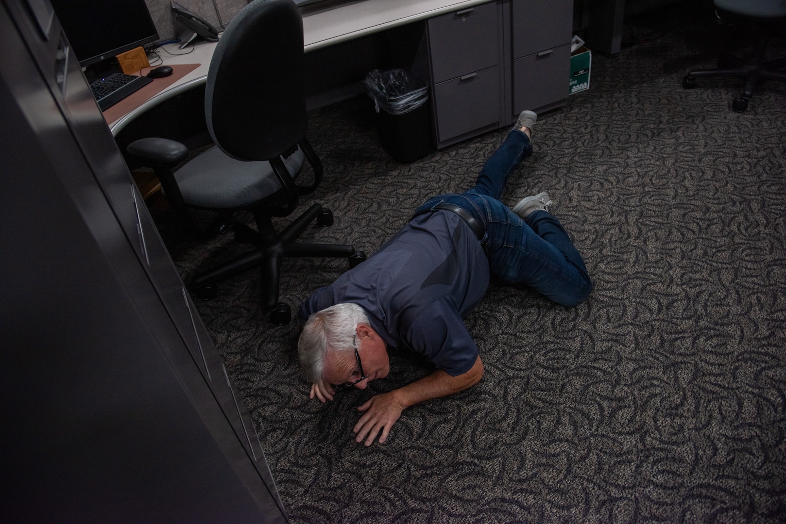 A man with gray hair wearing blue jeans and an blue shirt lays on floor behind desk and cabinets.