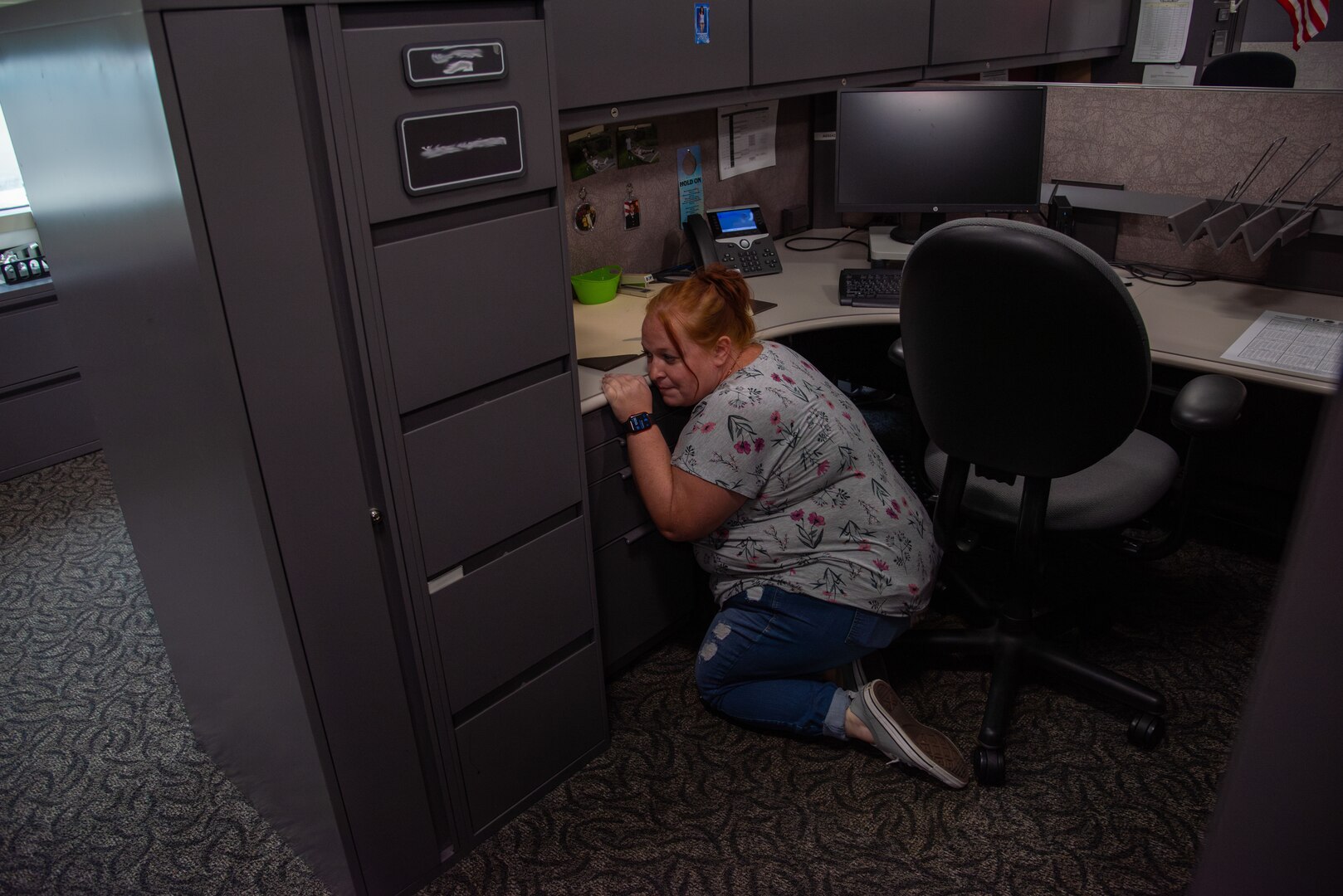 A woman wearing blue jeans and a gray shirt with flowers crouches behind a desk and cabinets.