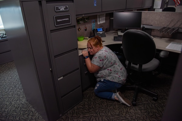 A woman wearing blue jeans and a gray shirt with flowers crouches behind a desk and cabinets.