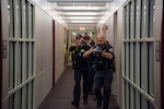 Police officers in uniform walk in a hallway toward camera, aiming blue replica training guns.