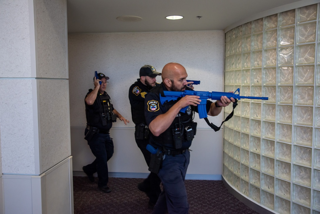 Three police officers in uniform walk around a glass block wall carrying blue replica training guns.