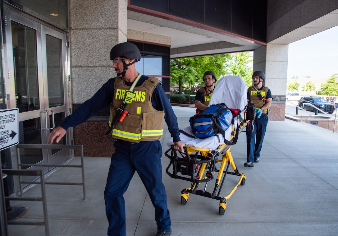 Three men wearing helmets, blue pants and tan and yellow FIRE/EMS vests pushing cot toward a building entrance.