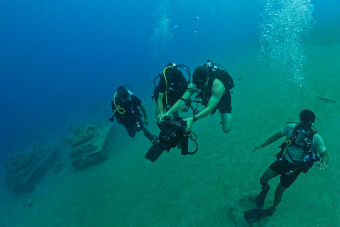 Service members wearing dive gear move underwater as one operates a piece of equipment.