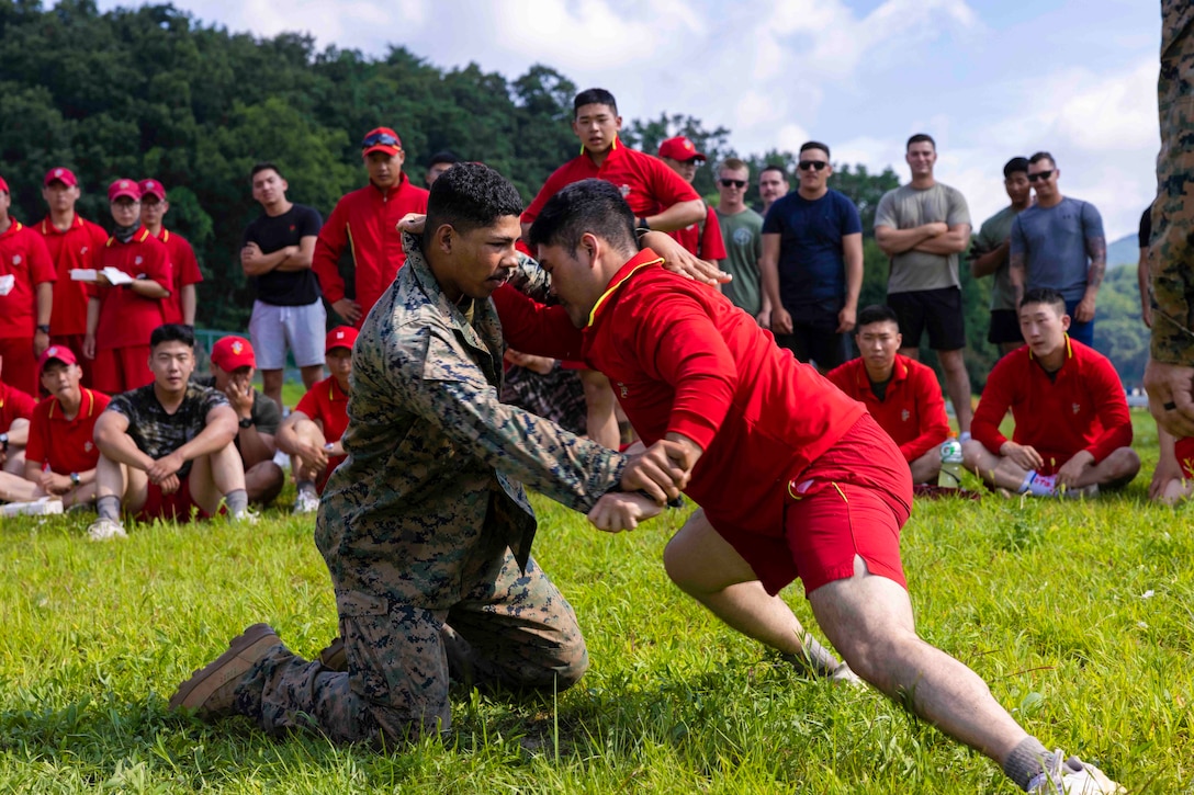 A U.S. Marine in uniform grapples with a South Korean service member wearing a red shirt and shorts as others watch.