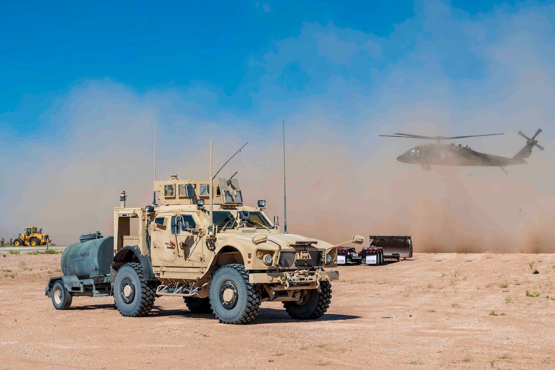 A military vehicle parked in front of an airborne helicopter.