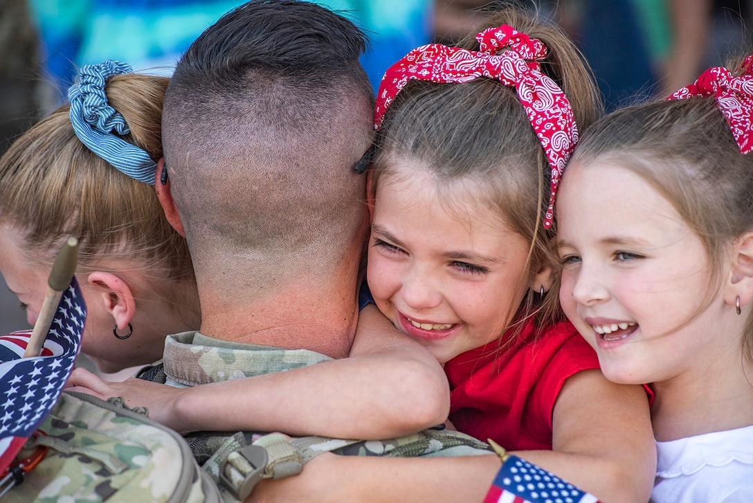 A soldier is photographed from behind and three smiling children hug the service member while holding American flags.