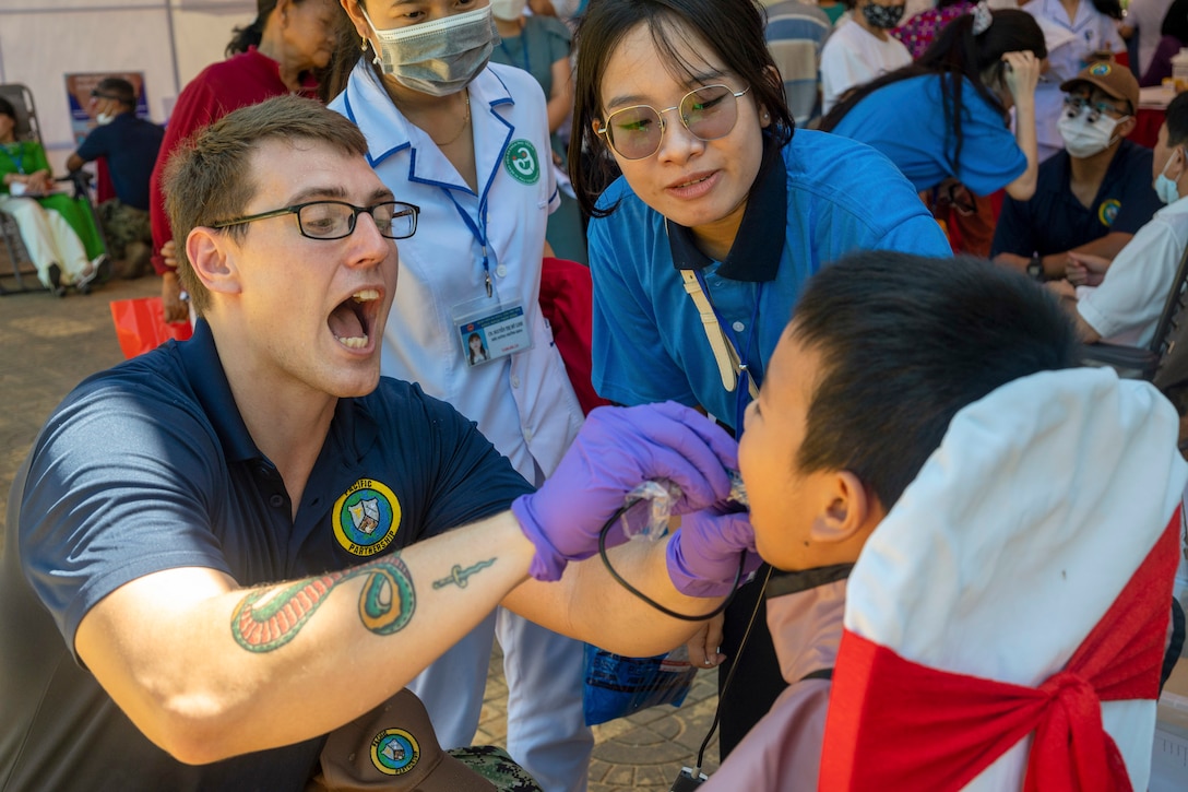 A sailor kneels while using a tool in a patient’s mouth as two people watch.