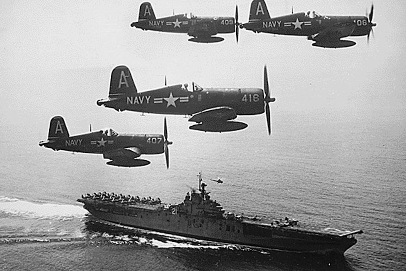 Four airplanes fly above an assault ship in the ocean.