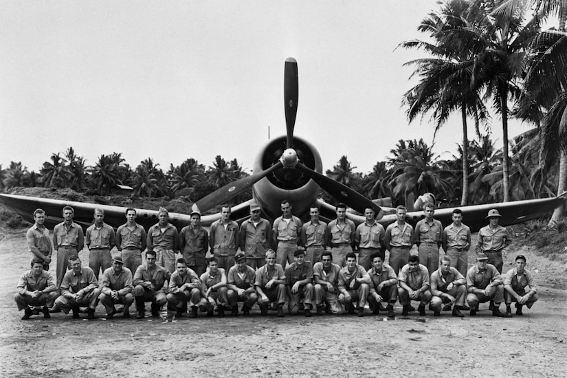 Two rows of service members stand in front of a single-engine propeller aircraft with palm trees in the background.