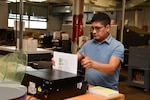 A man in a blue shirt stands at a work counter and feeds paper into a machine that sit on the counter.