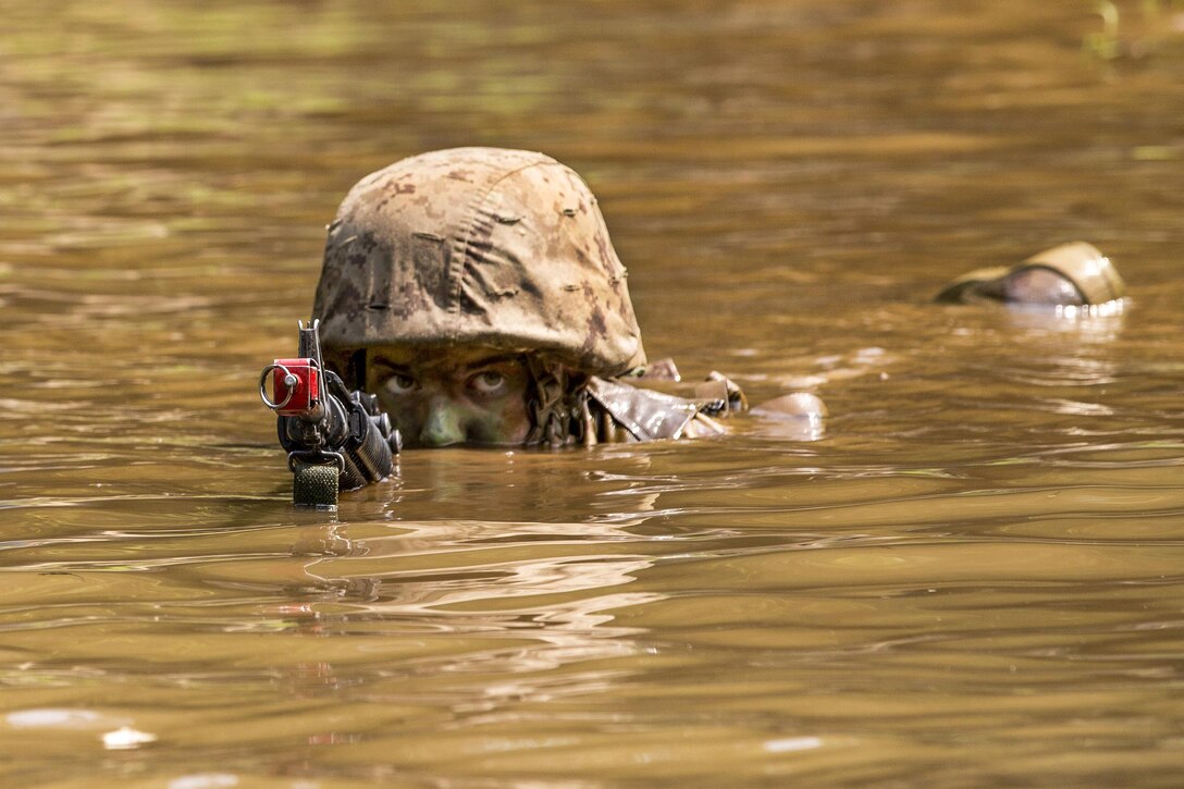 The head of a Marine can be seen from the nose up from muddy water. The eyes of the Marine can be seen and he is looking at the camera. his hands are under water but the barrel of his weapon is just in front of his face.