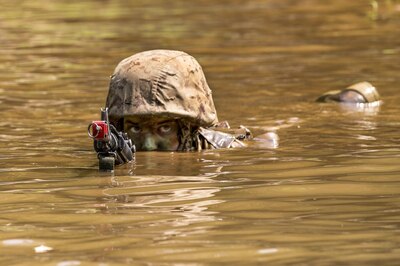 The head of a Marine can be seen from the nose up from muddy water. The eyes of the Marine can be seen and he is looking at the camera. his hands are under water but the barrel of his weapon is just in front of his face.
