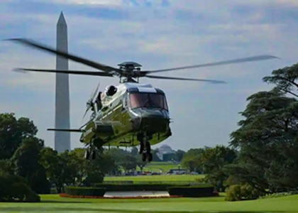 A green and white helicopter hovers just about the green grass. in the background is the Washington monument.