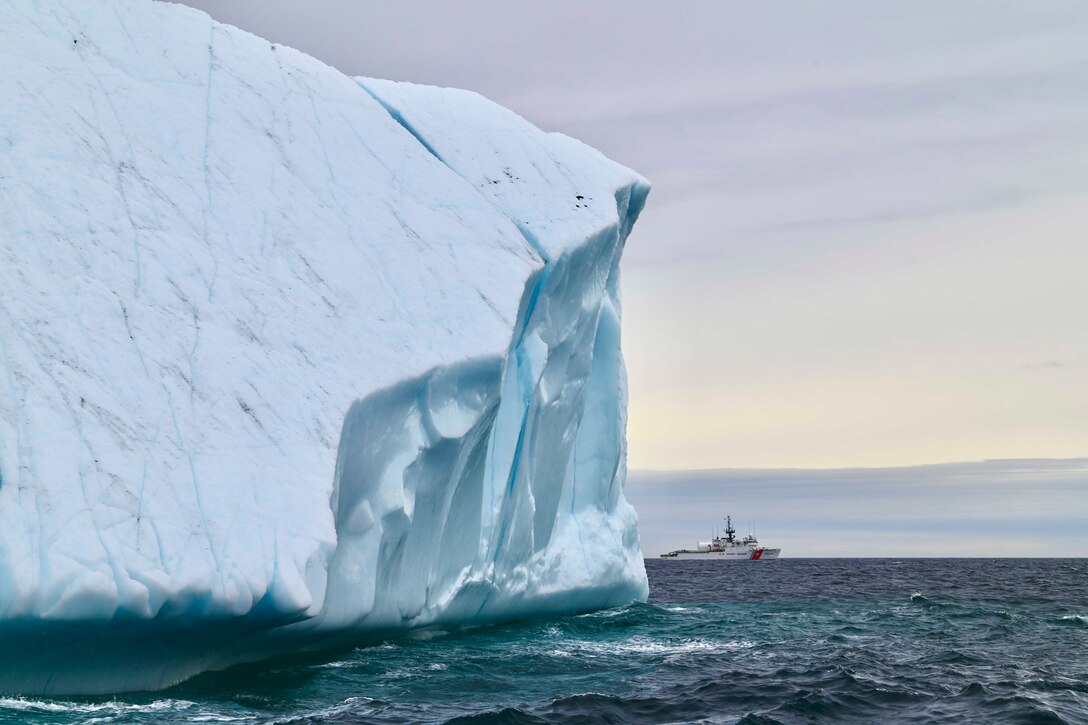 A boat travels through a body of water behind a large iceberg.