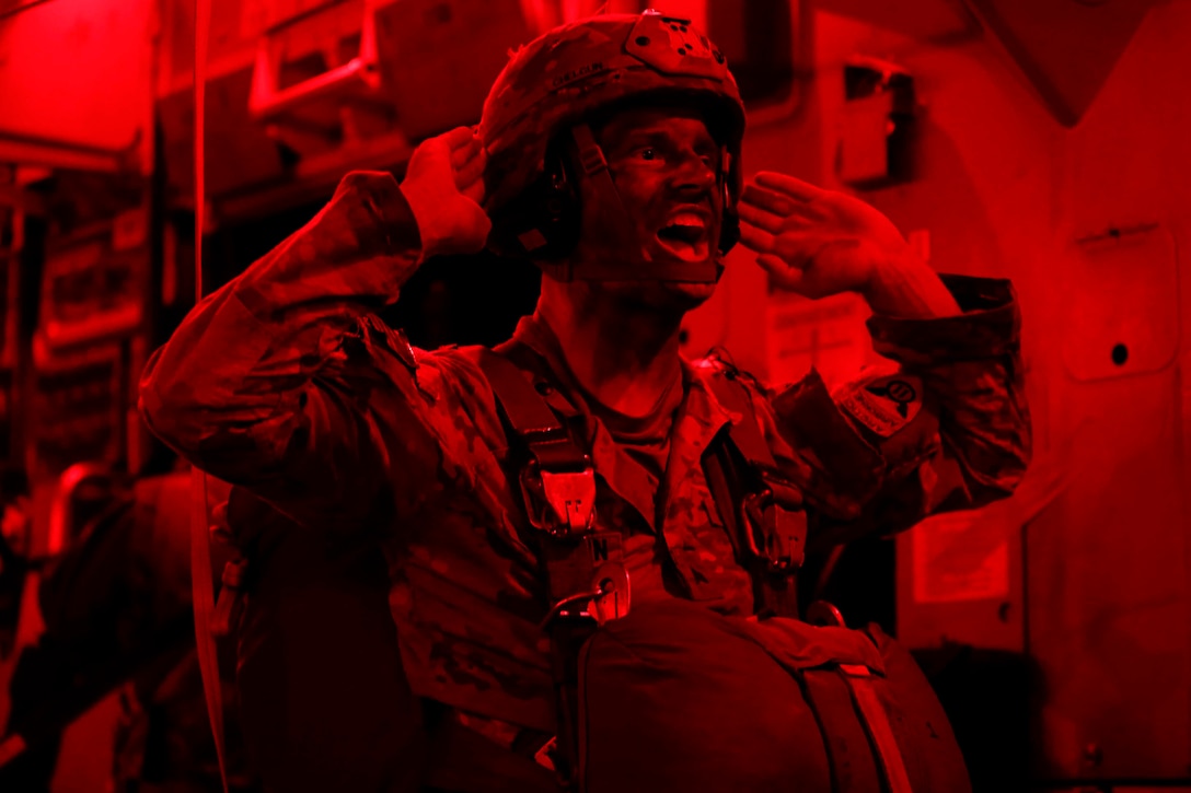 A soldier illuminated by red light holds his hands by his ears while speaking.