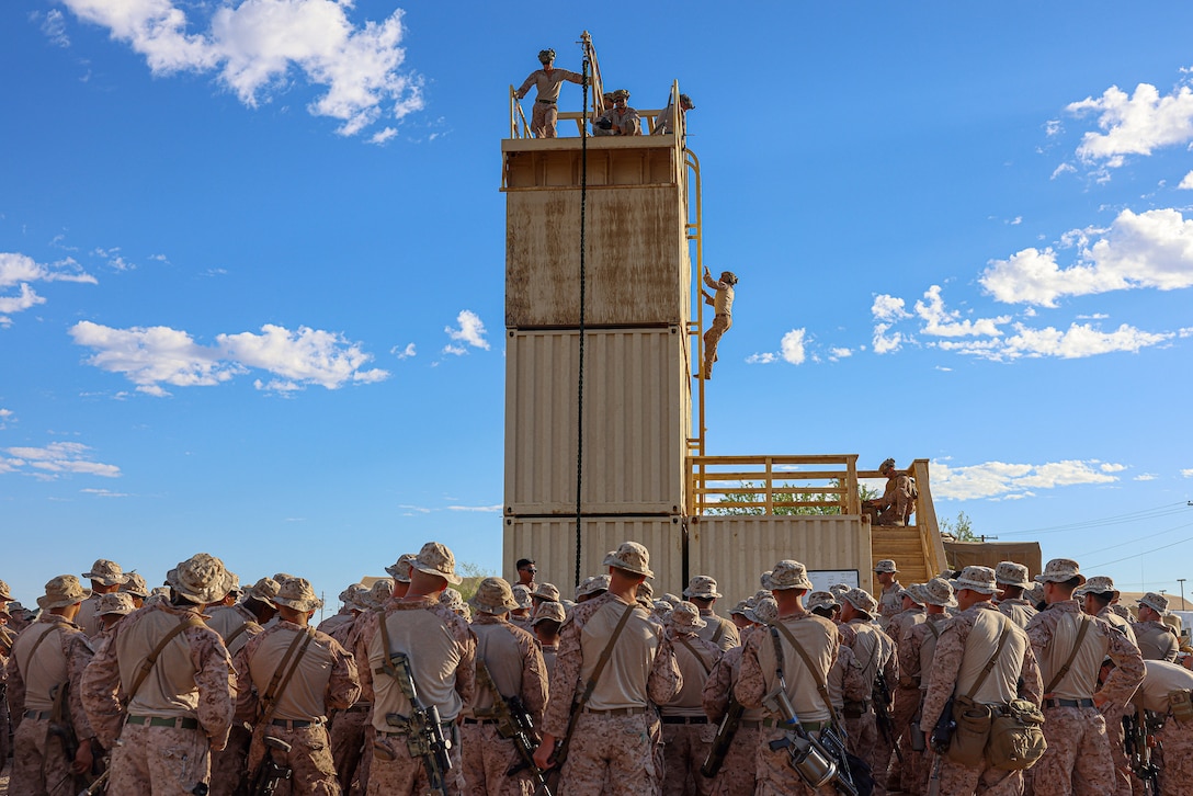 Marines crowd around the bottom of a fast-rope tower as one climbs it while others stand atop it.