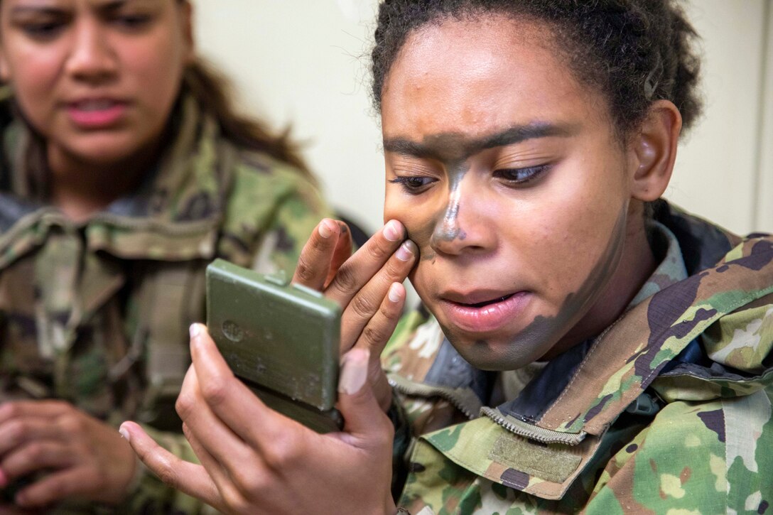 A soldier looks at a hand-held mirror while applying camouflage face paint as a fellow service member watches.