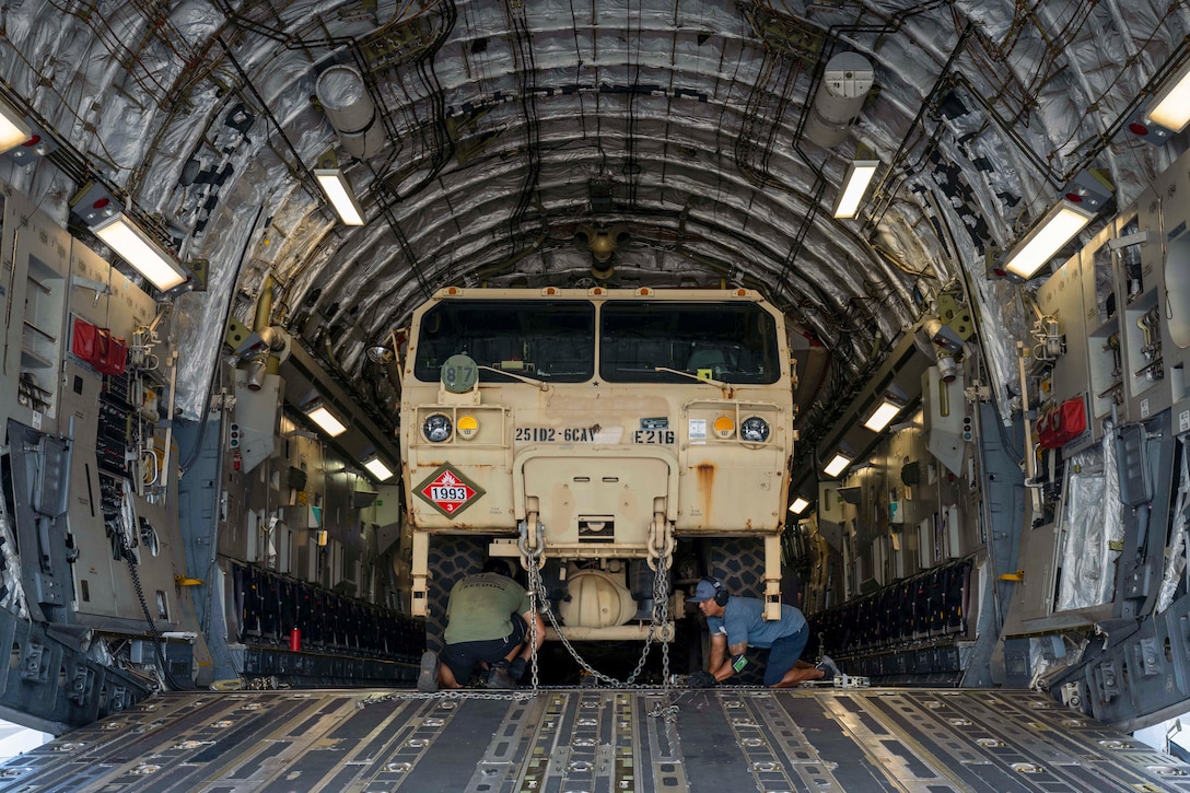 Two airmen kneel while securing a chain to a truck in an aircraft as seen through the open doorway.